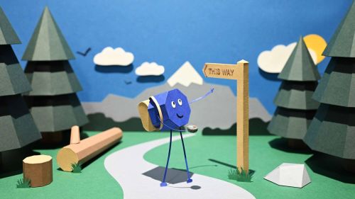 Paper-crafted character with a backpack and compass standing at a woodland path beside a signpost reading “This Way”, pointing towards distant mountains.