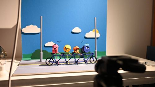 Behind-the-scenes photograph of a paper-crafted tandem bicycle with four colourful characters.