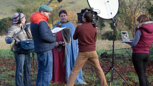 Behind-the-scenes view of a film crew shooting outdoors, with a costumed actor in medieval clothing, a camera operator, sound technician, lighting equipment, and other crew members.