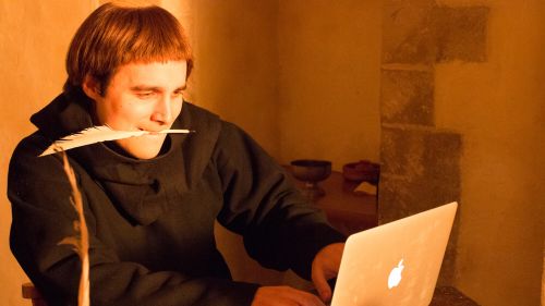 A man dressed in a dark medieval-style robe holds a feather quill in his mouth while using a laptop in a warmly lit stone interior.