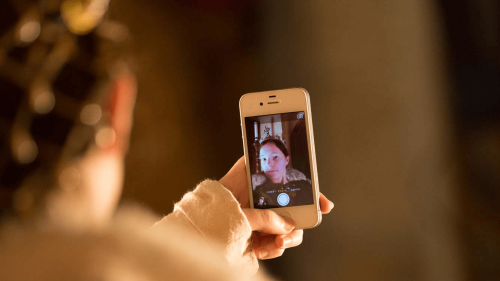 Over-the-shoulder view of a person holding a smartphone and photographing a woman displayed on the phone screen in warm indoor lighting.