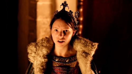 A woman wearing an ornate crown, fur-trimmed cloak, and medieval-style clothing looks toward the camera in a dim, warmly lit interior.