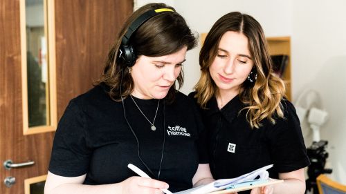 Two people reviewing notes together, with one wearing headphones and writing on a clipboard.